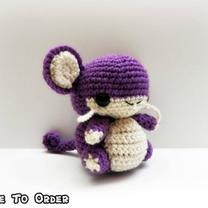 May include: A handmade crochet plush toy in the shape of a mouse. The mouse is primarily purple with cream-colored accents on the ears, belly, and feet. It has black eyes and white whiskers. The text "MADE TO ORDER" is at the bottom.