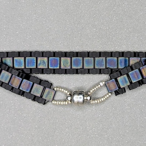 Op de afbeelding: Zwart kralenarmband met iriserende vierkante kralen. De armband heeft een magnetische sluiting en is ongeveer 18 cm lang.