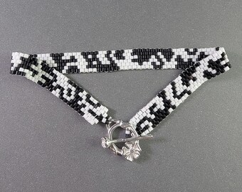 Puede incluir: Un collar choker de cuentas en blanco y negro con un patrón geométrico. El collar tiene un cierre plateado con un diseño floral. El choker se muestra sobre una superficie gris.