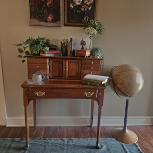 May include: A wooden secretary desk with a drop-down writing surface, multiple small drawers, and two central cabinet doors. The desk features brass hardware, a light-colored mug, books, and a potted plant on top. A floor globe stands to the right.