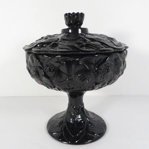 May include: A black, footed candy dish with a lid. The dish and lid are adorned with a floral relief pattern. The lid has a small, flower-shaped handle. The dish is made of a shiny, opaque material.