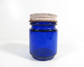 May include: A small, cylindrical cobalt blue glass jar with a threaded neck. The jar features a white metal screw-on lid with thin vertical red stripes and the printed text CAP MADE IN CANADA. The lid shows signs of oxidation and rust.