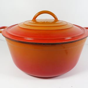 May include: A round, orange cast iron Dutch oven with a matching lid. The lid has a loop handle, and the pot has two side handles. The color gradient transitions from a deep red at the base to a bright orange at the top.