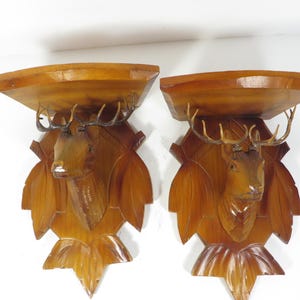 May include: Two wooden wall shelves with carved deer head and antler details. The shelves are a warm brown color and have a leaf-shaped design. The deer heads are intricately carved and add a rustic touch.