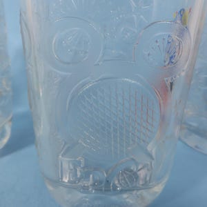 May include: Clear glass tumblers with embossed designs. One tumbler features a tennis racket and ball design, while others have floral and geometric patterns. The glasses are set against a light blue background.