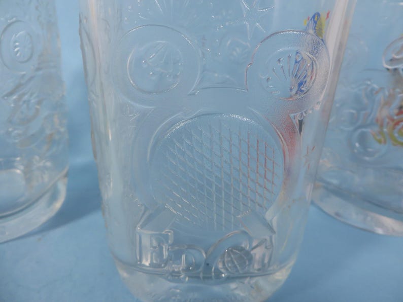 May include: Clear glass tumblers with embossed designs. One tumbler features a tennis racket and ball design, while others have floral and geometric patterns. The glasses are set against a light blue background.