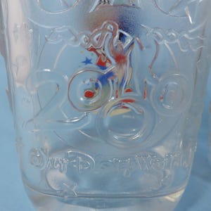 May include: Clear glass tumbler with embossed design. Features the number "2000" and the words "Disney World". Includes a colorful graphic of a character on a bicycle. Ideal for beverages.