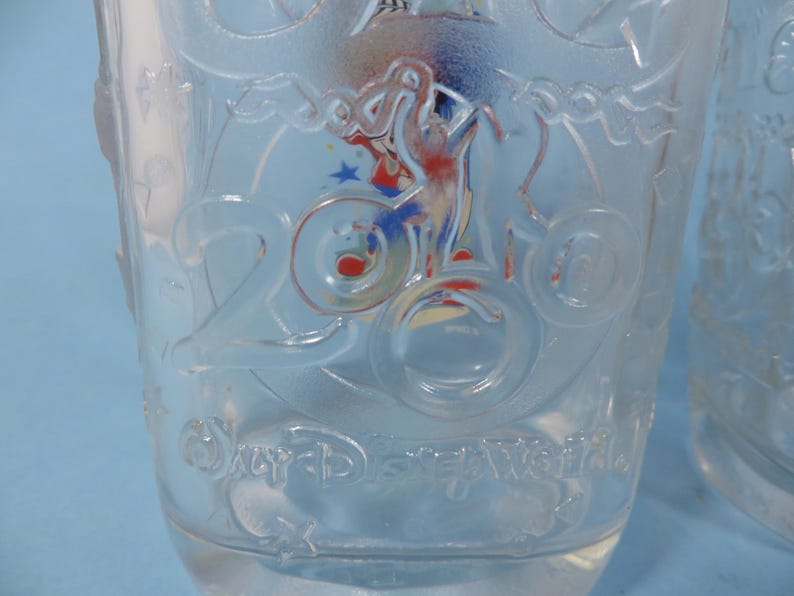 May include: Clear glass tumbler with embossed design. Features the number "2000" and the words "Disney World". Includes a colorful graphic of a character on a bicycle. Ideal for beverages.