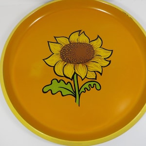 May include: A round, yellow serving tray with a vibrant sunflower design. The sunflower has yellow petals, a brown center, and green leaves and stem. The tray's rim is a lighter shade of yellow.