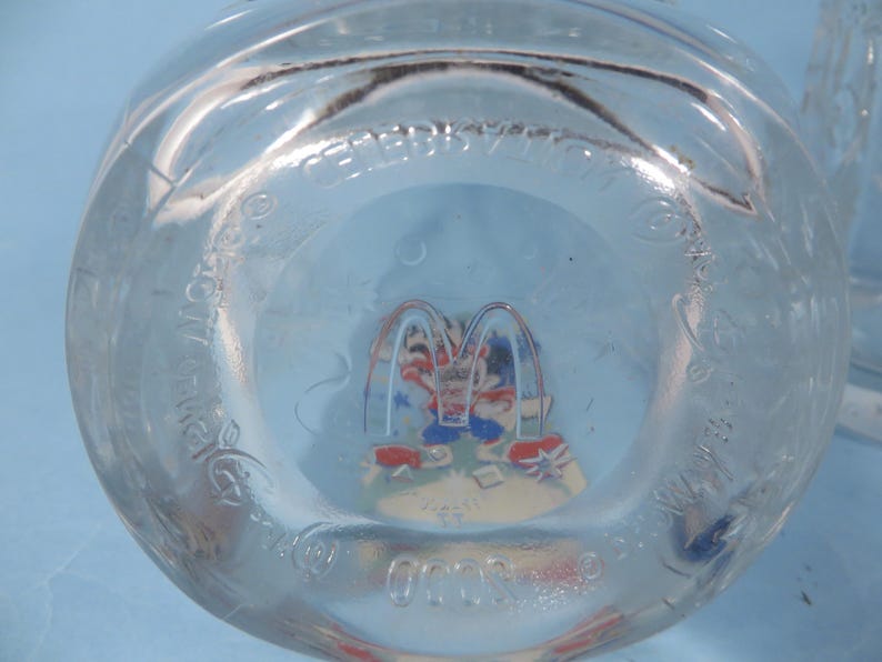 May include: Clear glass McDonald's cup with a colorful graphic of Ronald McDonald and friends. The cup has the McDonald's logo and text around the rim. The bottom of the cup features a colorful image of the McDonald's characters.