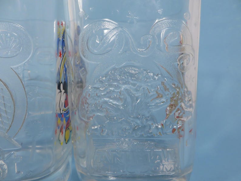 May include: Two clear glass tumblers with embossed designs. One features a stylized tree and the text "Walt Disney World". The other has a Mickey Mouse design. The glasses are on a light blue background.