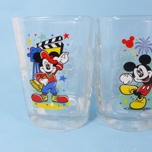 May include: Two clear drinking glasses featuring Mickey Mouse. One glass shows Mickey with a film clapper and camera, the other with fireworks. Both glasses have colorful graphics and the Disney logo.