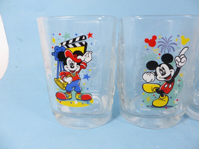 May include: Two clear drinking glasses featuring Mickey Mouse. One glass shows Mickey with a film clapper and camera, the other with fireworks. Both glasses have colorful graphics and the Disney logo.