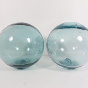 May include: Two spherical glass floats in a translucent light blue color. Each orb features a smooth, rounded surface with subtle internal air bubbles and a small, circular glass seal mark at the base, reflecting light on their curved surfaces.