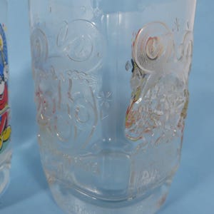 May include: Clear glass drinking glasses with embossed Disney character designs. One glass features Mickey Mouse with fireworks, the other has a detailed embossed design. The glasses are suitable for cold beverages.