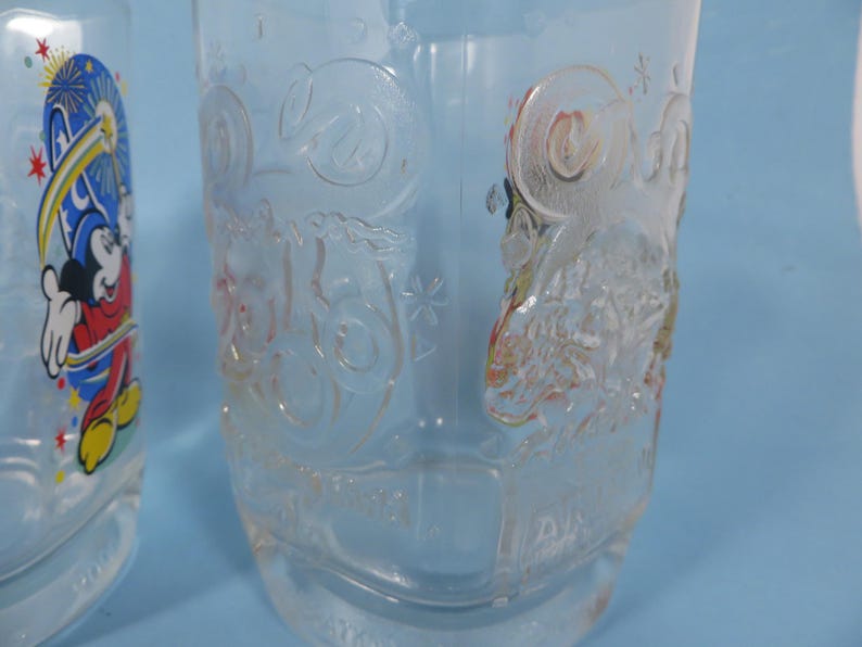 May include: Clear glass drinking glasses with embossed Disney character designs. One glass features Mickey Mouse with fireworks, the other has a detailed embossed design. The glasses are suitable for cold beverages.