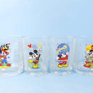 May include: Four clear drinking glasses with colorful Mickey Mouse designs. Each glass features a different pose of Mickey Mouse, with various backgrounds and accents. The glasses are set against a light blue background.