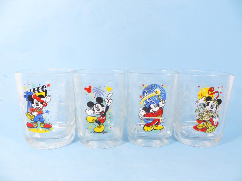 May include: Four clear drinking glasses with colorful Mickey Mouse designs. Each glass features a different pose of Mickey Mouse, with various backgrounds and accents. The glasses are set against a light blue background.