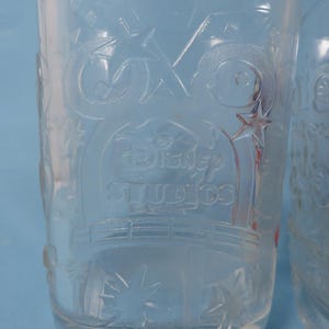 May include: Clear glass tumbler with embossed Disney Studios logo and star accents. The glass has a cylindrical shape and is partially filled. The design is visible against a light blue background.