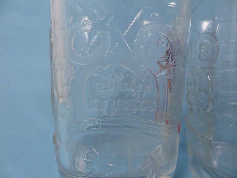 May include: Clear glass tumbler with embossed Disney Studios logo and star accents. The glass has a cylindrical shape and is partially filled. The design is visible against a light blue background.