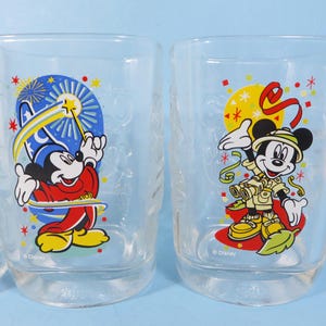 May include: Two clear drinking glasses with colorful Disney character designs. One features Mickey Mouse in a magician's outfit with a wand, the other shows Mickey in safari attire. Both glasses have a clear base.