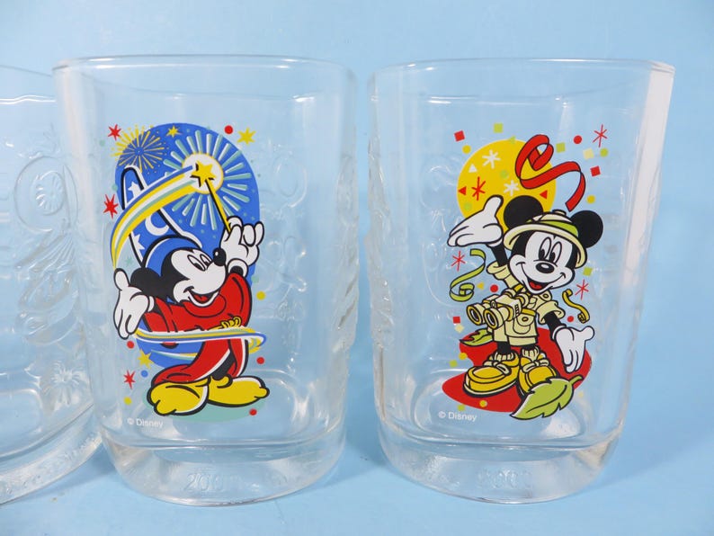 May include: Two clear drinking glasses with colorful Disney character designs. One features Mickey Mouse in a magician's outfit with a wand, the other shows Mickey in safari attire. Both glasses have a clear base.