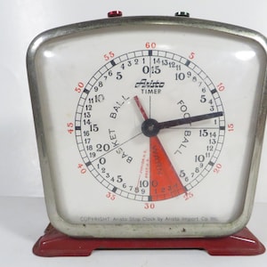 May include: A silver and red vintage Aristo timer with a white face. The face has three concentric circles with markings for basketball, football, and warning. The timer has a black hand and a red hand.