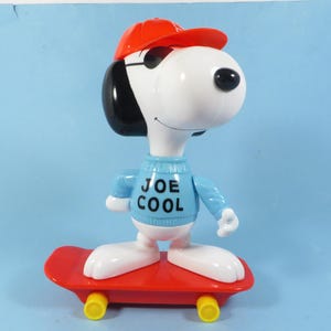 May include: A Snoopy figurine on a red skateboard, wearing a red cap and blue sweater with "JOE COOL" text. The skateboard has yellow wheels. The figurine is white, black, and red.