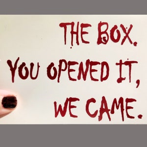 Op de afbeelding: Een witte kaart met de woorden "THE BOX. YOU OPENED IT, WE CAME." geschreven in rode inkt. De tekst heeft een druipend effect, wat een horrorthema suggereert. Een vingernagel met donkere nagellak is zichtbaar in de linker benedenhoek.