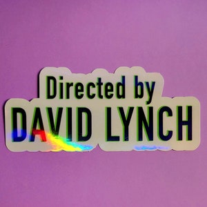 Op de afbeelding: Een sticker met de tekst "Directed by DAVID LYNCH" in groene en zwarte letters. De sticker heeft een holografische, regenboogkleurige glans. De sticker staat op een paarse achtergrond.