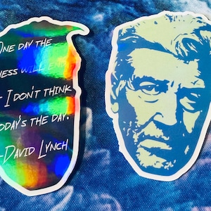 Puede incluir: Dos pegatinas, una con fondo holográfico y la otra con una imagen azul y blanca de la cara de un hombre. La pegatina holográfica tiene una cita de David Lynch que dice: "Un día la tristeza terminará, pero no creo que sea hoy."