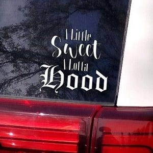 Puede incluir: Una calcomanía de vinilo blanco con el texto "A Little Sweet A Lotta Hood" sobre un fondo negro. La calcomanía está aplicada en la ventana trasera de un coche.