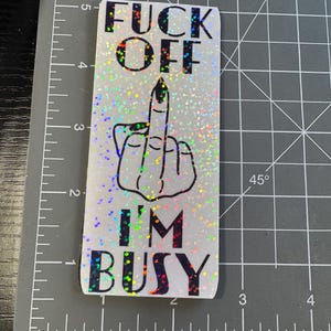 May include: A white sticker with a glittery finish and black text that reads "F**k Off I'm Busy". The sticker features a black and white illustration of a middle finger.