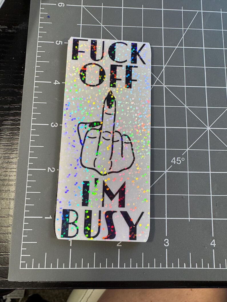 May include: A white sticker with a glittery finish and black text that reads "F**k Off I'm Busy". The sticker features a black and white illustration of a middle finger.