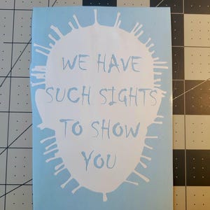 Op de afbeelding: Lichtblauwe sticker met witte tekst: "WE HAVE SUCH SIGHTS TO SHOW YOU". De tekst staat in een witte vorm met een spiky, zon-achtig ontwerp eromheen.