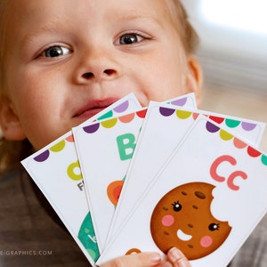 May include: A child holds up colorful alphabet flash cards with letters "B", "C", and "F" printed on them. The "C" card features a cartoon cookie with a bite taken out of it.