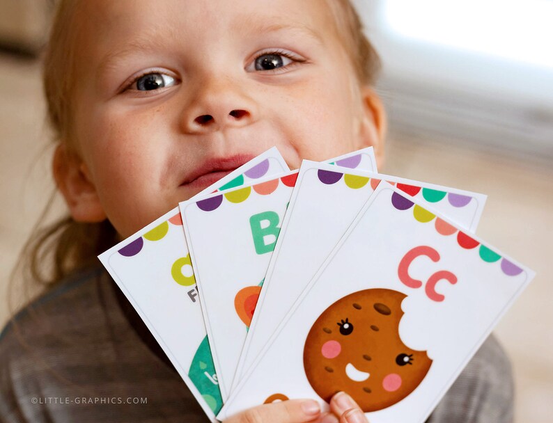 May include: A child holds up colorful alphabet flash cards with letters "B", "C", and "F" printed on them. The "C" card features a cartoon cookie with a bite taken out of it.