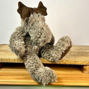 May include: A plush, seated stuffed animal with brown fur and a dark brown bow on its head. The toy has long, shaggy fur and is positioned on a wooden surface. The toy's feet and bow are a darker shade of brown.