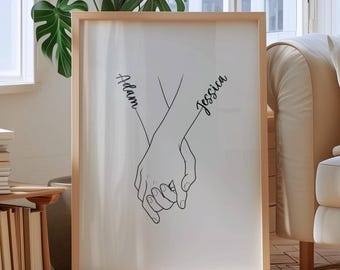 May include: A framed art print featuring a minimalist line drawing of two hands holding each other. The names "Adam" and "Jessica" are written in cursive on the arms. The artwork is displayed in a light-colored wooden frame, creating a simple and elegant aesthetic.