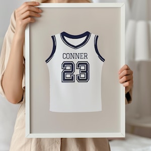 Könnte beinhalten: Gerahmter Druck mit einem weißen Basketballtrikot-Design mit marineblauen Akzenten. Das Trikot zeigt den Namen "CONNER" und die Nummer "23" in Marineblau. Der Rahmen ist weiß.