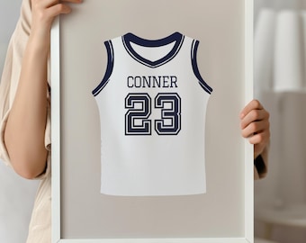 May include: A framed print featuring a white basketball jersey design with navy blue accents. The jersey displays the name "CONNER" and the number "23" in navy blue. The frame is white.