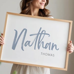 Op de afbeelding: Een rechthoekig, ingelijst bord met de naam "Nathan" in een lichtblauw scriptlettertype en de naam "Thomas" in een kleiner, zwart, schreefloos lettertype. Het frame is gemaakt van lichtgekleurd hout. De achtergrond is wit.