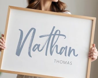 May include: A framed rectangular sign with the name "Nathan" in a light blue script font, and the name "Thomas" in a smaller, black, sans-serif font. The frame is made of light-colored wood. The background is white.