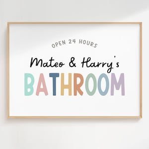 May include: A framed print with the words "Open 24 Hours" above "Mateo & Harry's BATHROOM" in colorful letters. The print has a light wood frame and is displayed on a white wall.