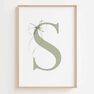 Op de afbeelding: Een ingelijste print met de letter "S" in een zachte saliegroene kleur. Een delicaat strikdesign is kunstzinnig boven de letter geplaatst, met vloeiende lijnen die een elegante touch toevoegen. Het kunstwerk staat tegen een witte achtergrond.