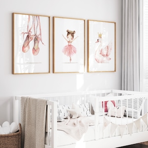 Puede incluir: Tres impresiones enmarcadas con ilustraciones de acuarela de zapatillas de ballet, una bailarina y un cisne. Las impresiones están colgadas en una pared blanca sobre una cuna blanca con una manta y almohadas blancas.