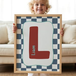 May include: A framed print displaying a large, brick-red letter "L" with the name "Liam" in teal. The letter is set against a white background with a teal border, framed by a blue and white checkered pattern. The frame is made of light-coloured wood.