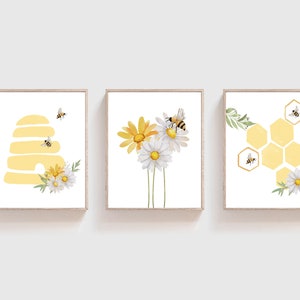 Op de afbeelding: Drie ingelijste prints met een bij- en bloementhema. De eerste print toont een gele honingraat met een bij die er voor vliegt. De tweede print toont een bij die vliegt in de buurt van een tros witte en gele madeliefjes. De derde print toont een honingraatpatroon met een bij die er in de buurt vliegt.