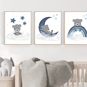 Op de afbeelding: Drie ingelijste prints met een grijze beer op een wolk met een blauwe halve maan, sterren en een regenboog. De prints zijn perfect voor een kinderkamer of babykamer.
