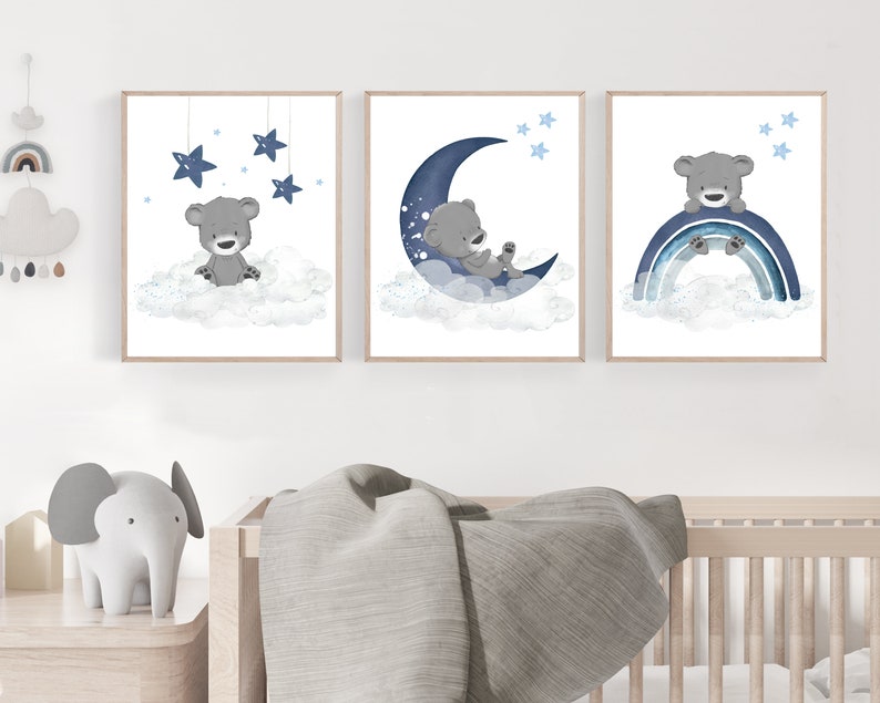 Op de afbeelding: Drie ingelijste prints met een grijze beer op een wolk met een blauwe halve maan, sterren en een regenboog. De prints zijn perfect voor een kinderkamer of babykamer.